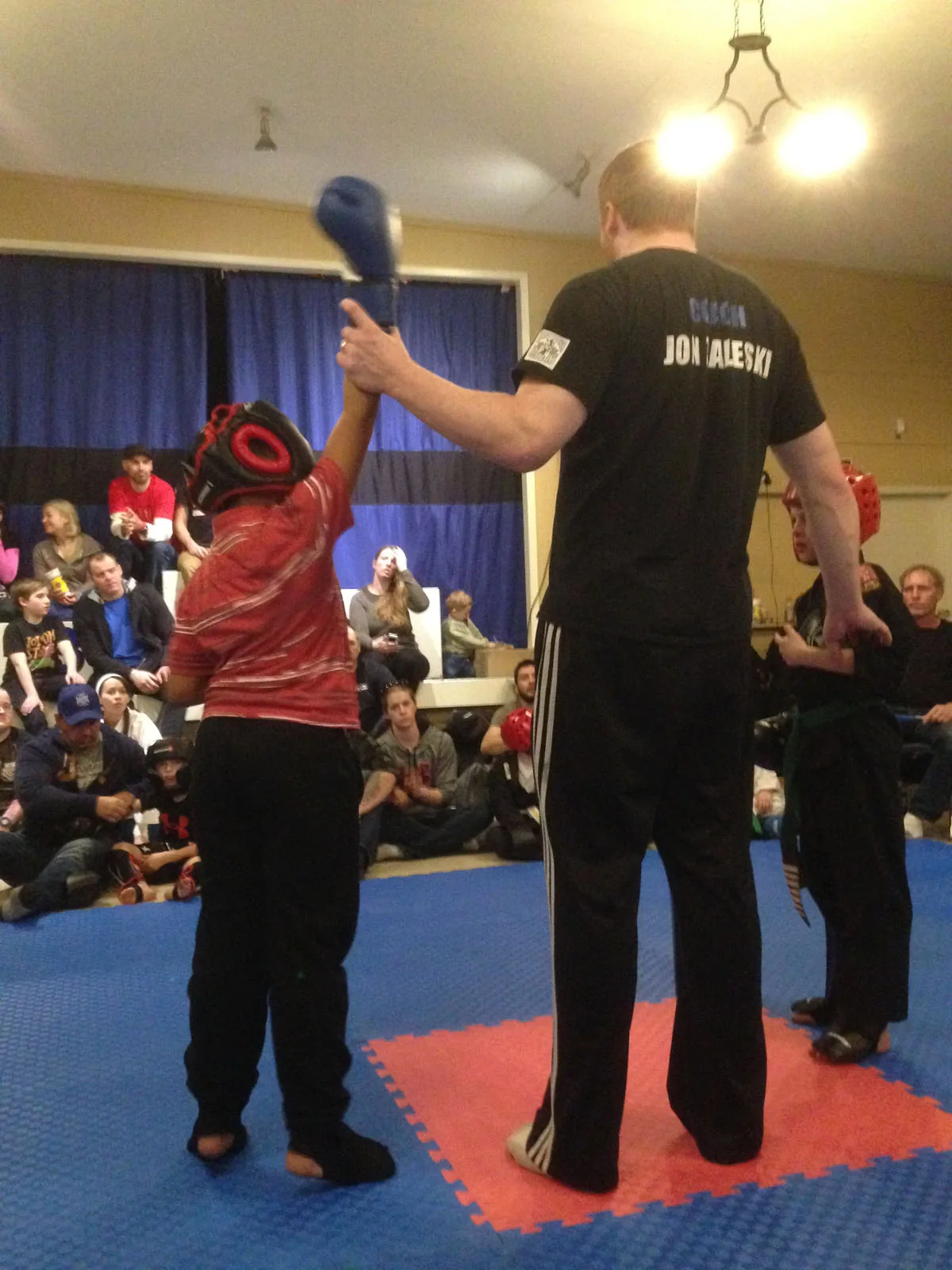 A coach raises a young student's glove after a kickboxing round while families watch from the mats